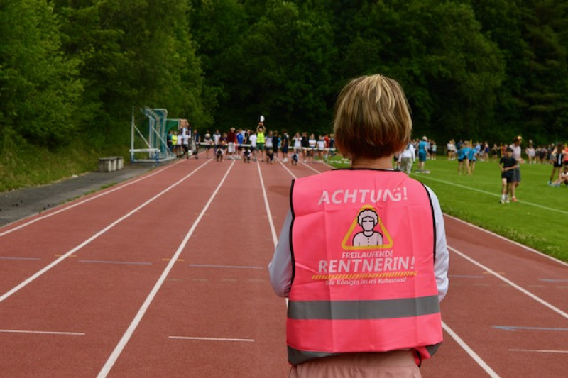 Frau mit Warnweste "Achtung! Freilaufende Rentnerin" auf Laufbahn bei Sportveranstaltung
